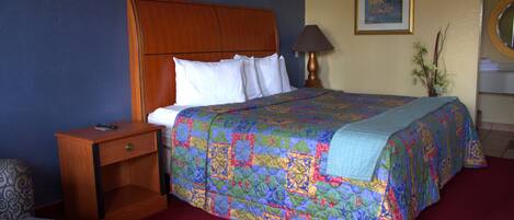 A hotel room with a wooden headboard, a bed with a colorful quilt, a bedside table with a lamp, and a chair.