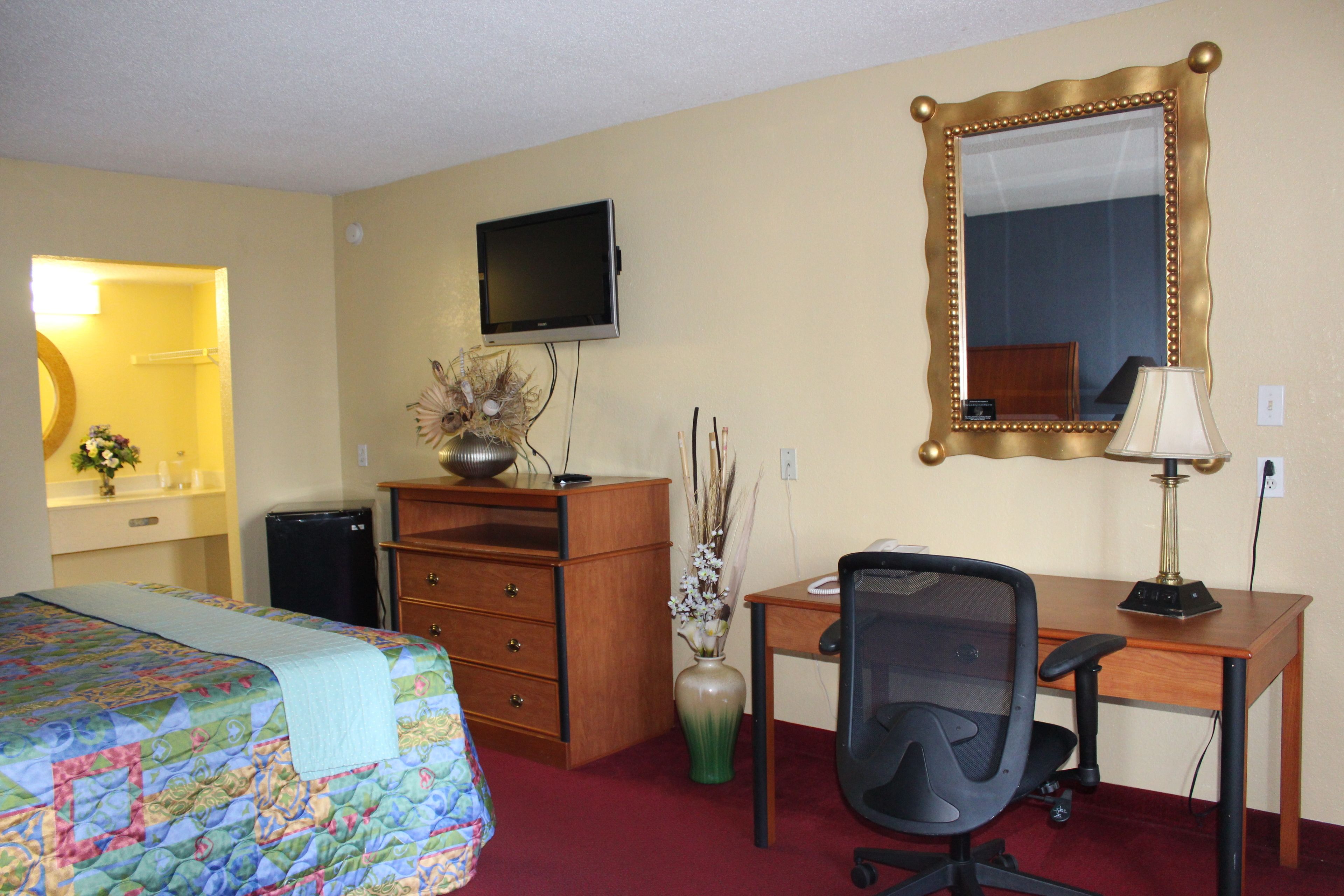 A hotel room with a bed, a desk with a chair, a television, a mirror, and a vase with flowers.