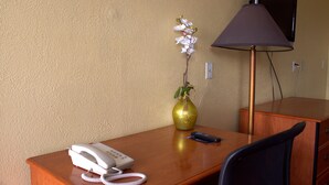 A hotel room with a wooden desk, a white telephone, a remote control, a vase with flowers, a lamp, and a television on a stand. - Lambert Inn (Kissimmee)