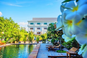 A pool area with lounge chairs and potted plants, a large building in the background, and a clear blue sky.