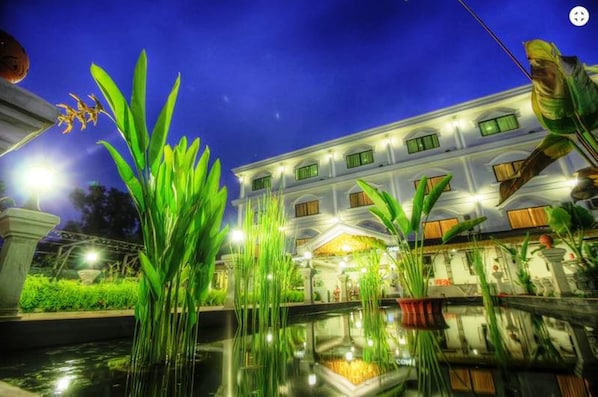 A well-lit hotel with a large pool, surrounded by lush greenery and a night sky.