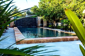 Une piscine rectangulaire bordée de pierres, entourée de verdure et d’un mur de pierres.