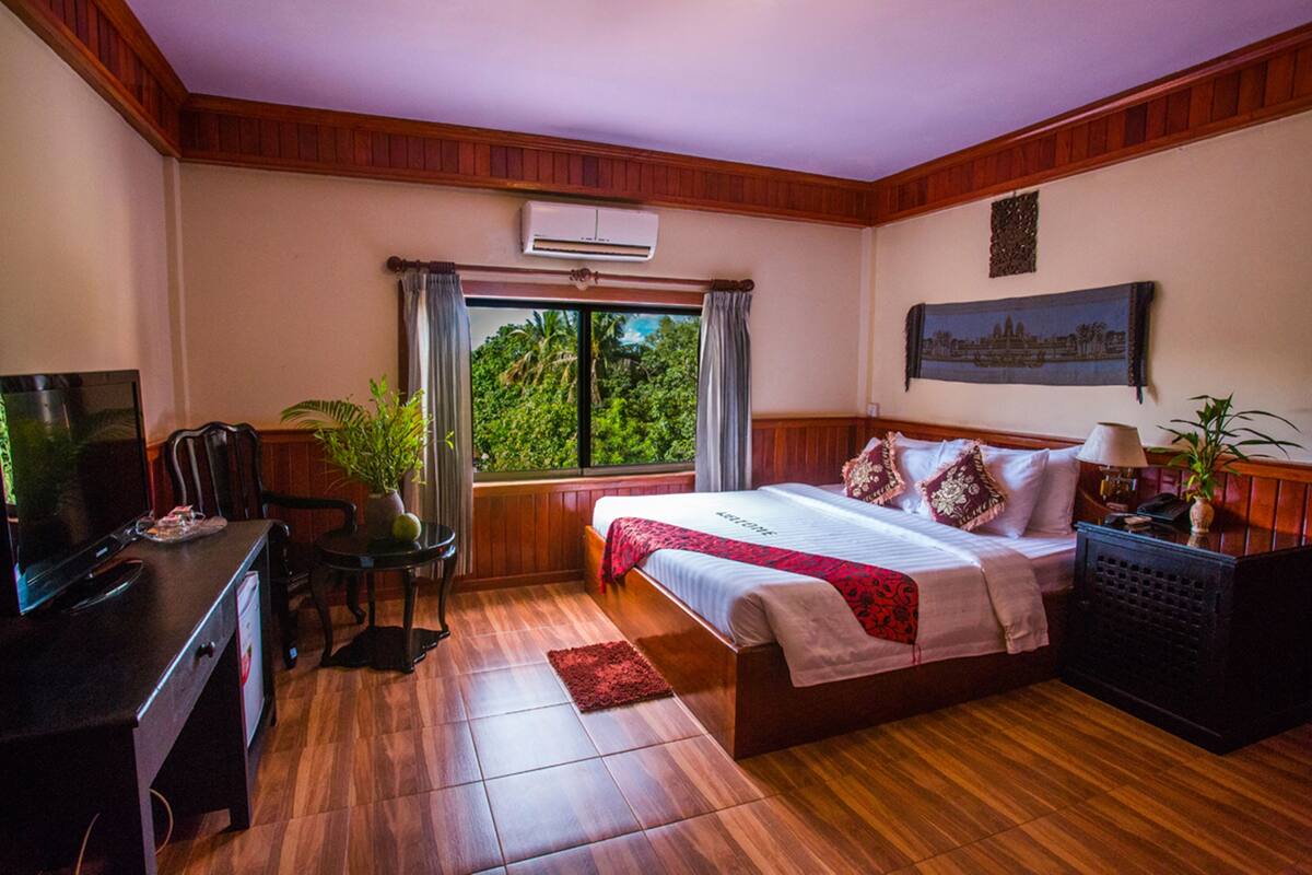 Een hotelkamer met een houten vloer, een bed met een rood-witte dekbedovertrek, een bureau met een televisie, een stoel en een raam met gordijnen.