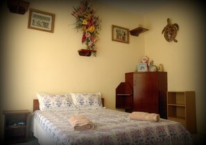 A bedroom with a bed, a wooden wardrobe, a small shelf, and a wall-mounted decoration. - Reve des Iles (Rodrigues Island)