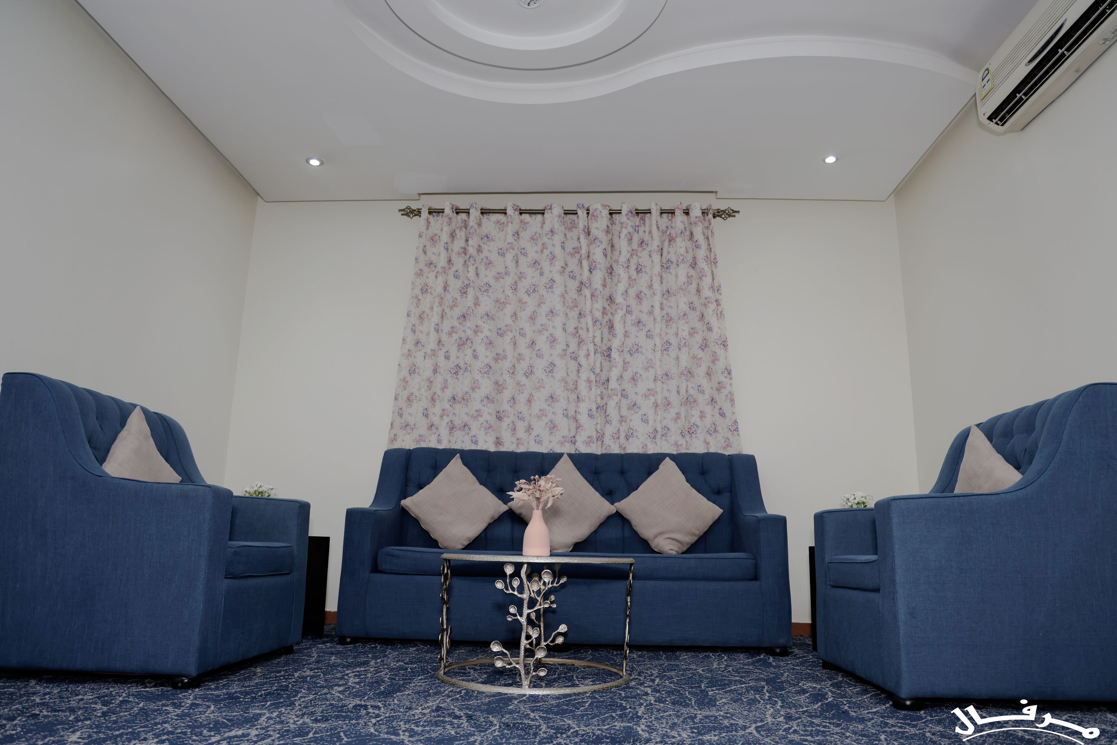 Een kamer met een blauwe bank, twee fauteuils, een tafeltje met een vaas, en een kamerbreed tapijt met dessin.