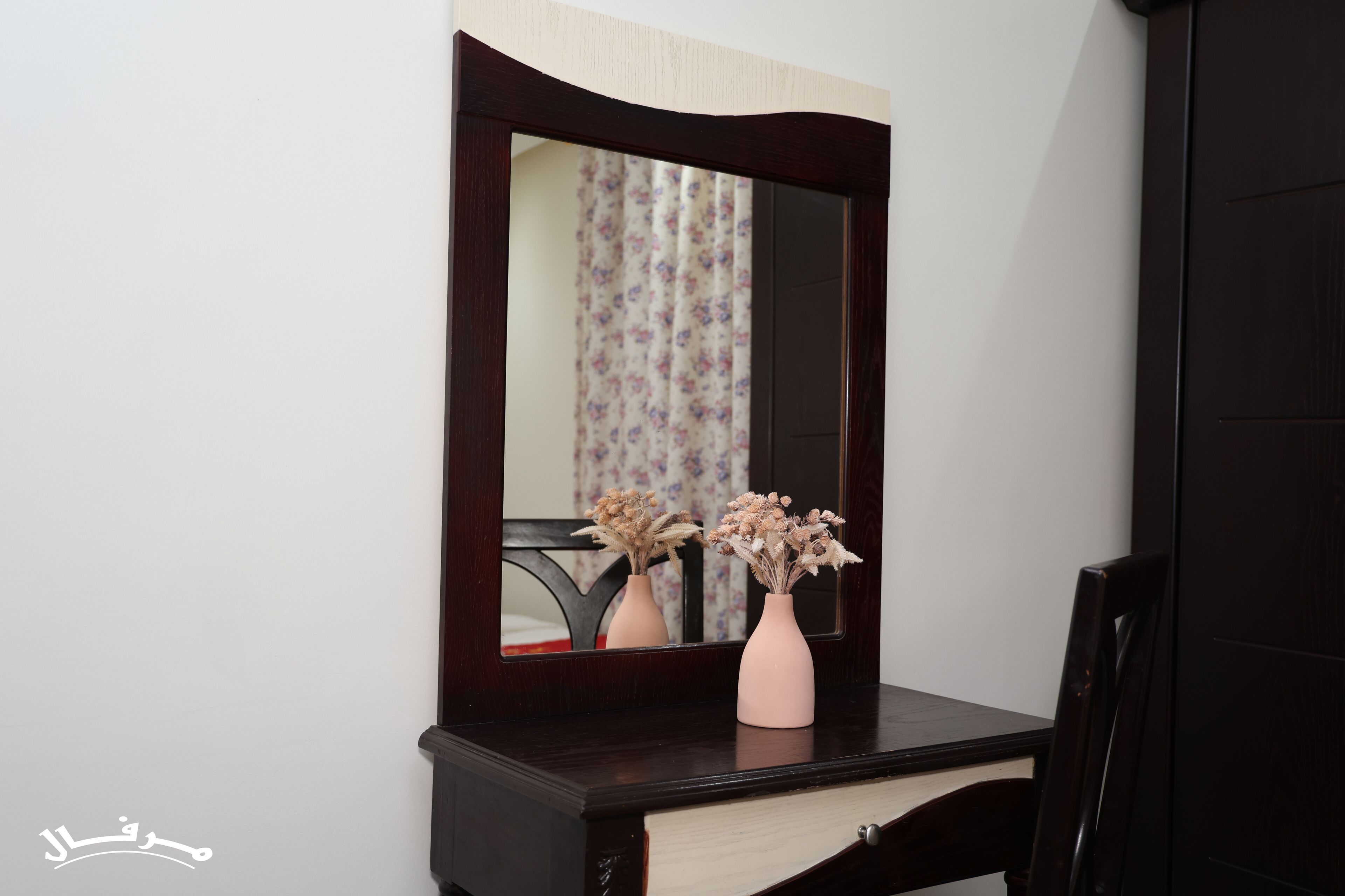 A dark wooden dresser with a mirror above it, two pink vases with dried flowers, and a floral curtain in the background.