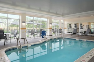Una piscina interior con paredes de vidrio transparente, borde azul y una zona de estar con mesas y sillas.