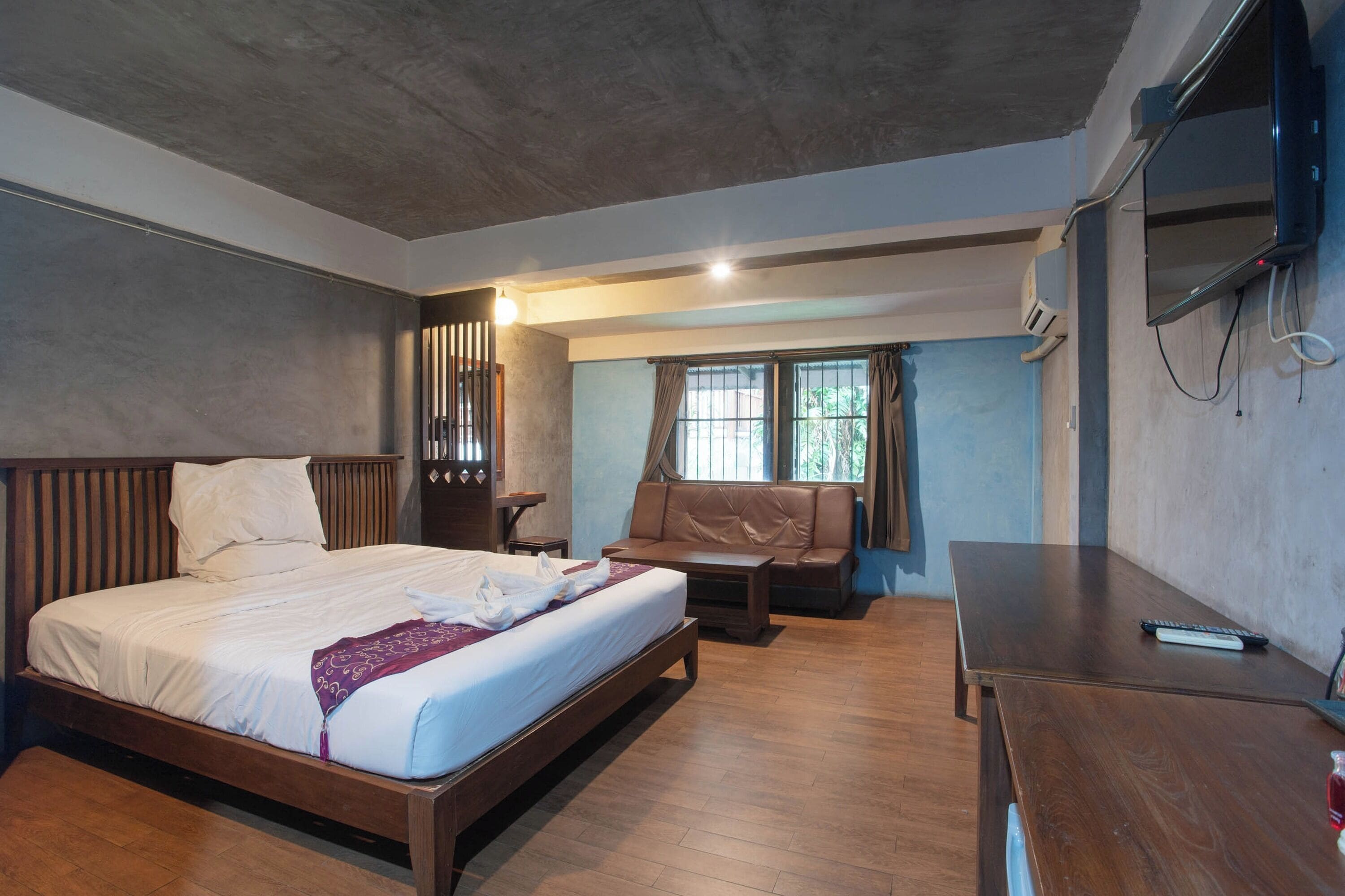 A modern bedroom with a wooden bed, a sofa, a desk, and a TV mounted on the wall.