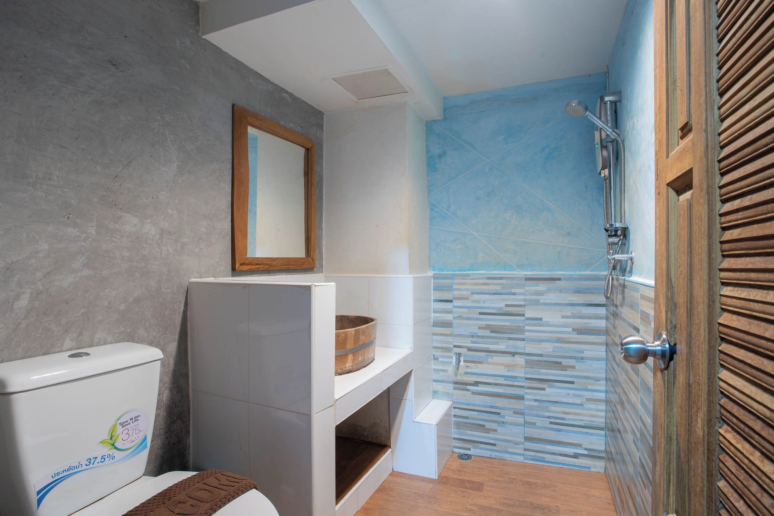 Un baño con inodoro, estante de madera, espejo y zona de ducha con pared de azulejos.
