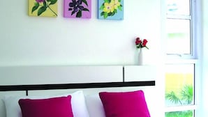 A hotel room with a bed, purple and white bedding, a window with a view of greenery, and three floral paintings on the wall. - Baan Rim Klong Resort (Sukhothai)