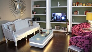 A living room with a white sofa, a glass coffee table, a pink patterned sofa, a television, and shelves with various items. - Andromeda Suites and Apartments (Athens)