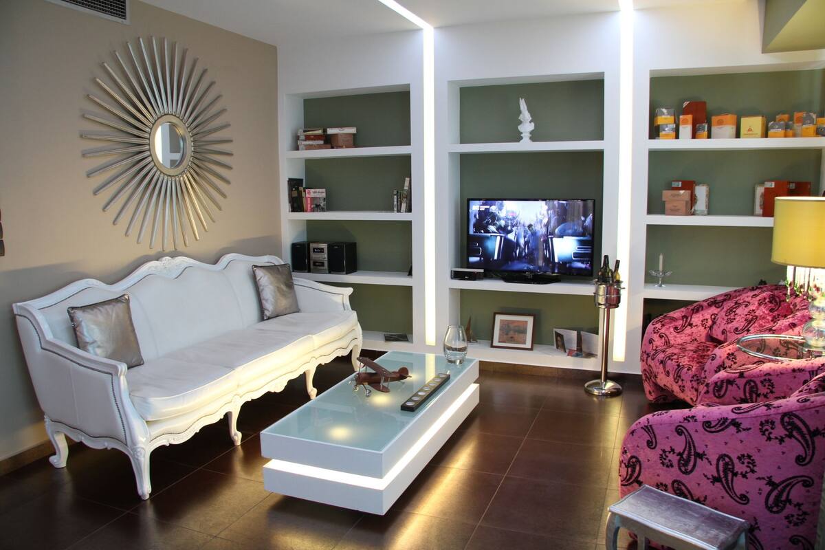 A living room with a white sofa, a glass coffee table, a pink patterned sofa, a television, and shelves with various items.