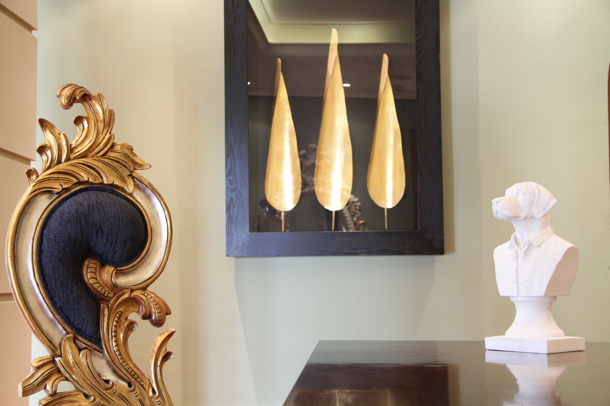 A room with a dark wooden table, a gold ornate chair, a white bust, and three tall, slender light fixtures on the wall.