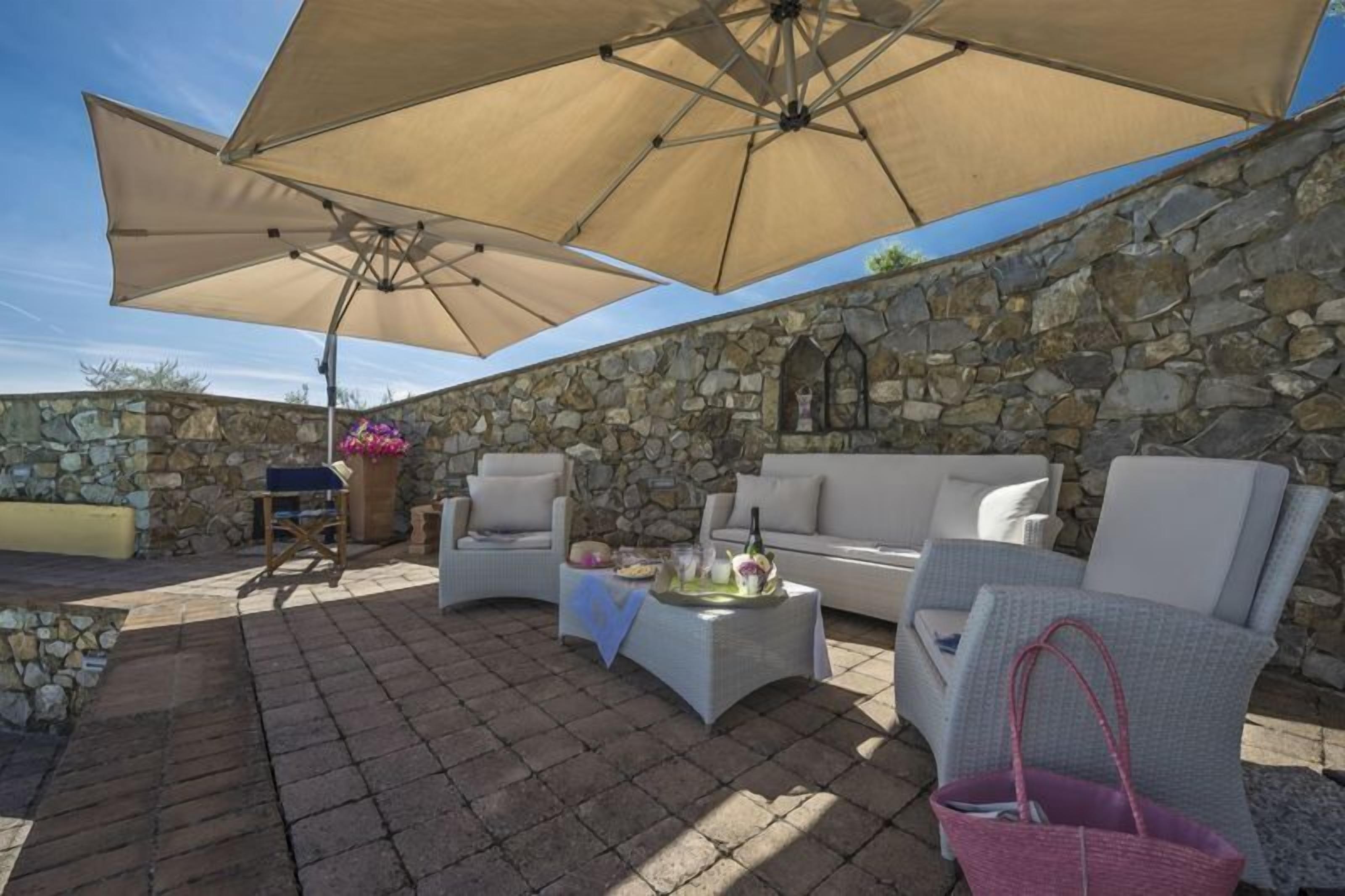 Eine Terrassengaststätte mit Steinmauern, einem weißen Sofa, zwei Sesseln, einem Couchtisch und großen, beigefarbenen Sonnenschirmen, die Schatten spenden.