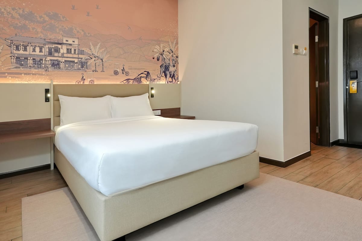 A neatly made bed in a hotel room with a large wall mural, a wooden floor, and a door to the right.