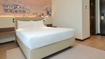 A neatly made bed in a hotel room with a large wall mural, a wooden floor, and a door to the right.