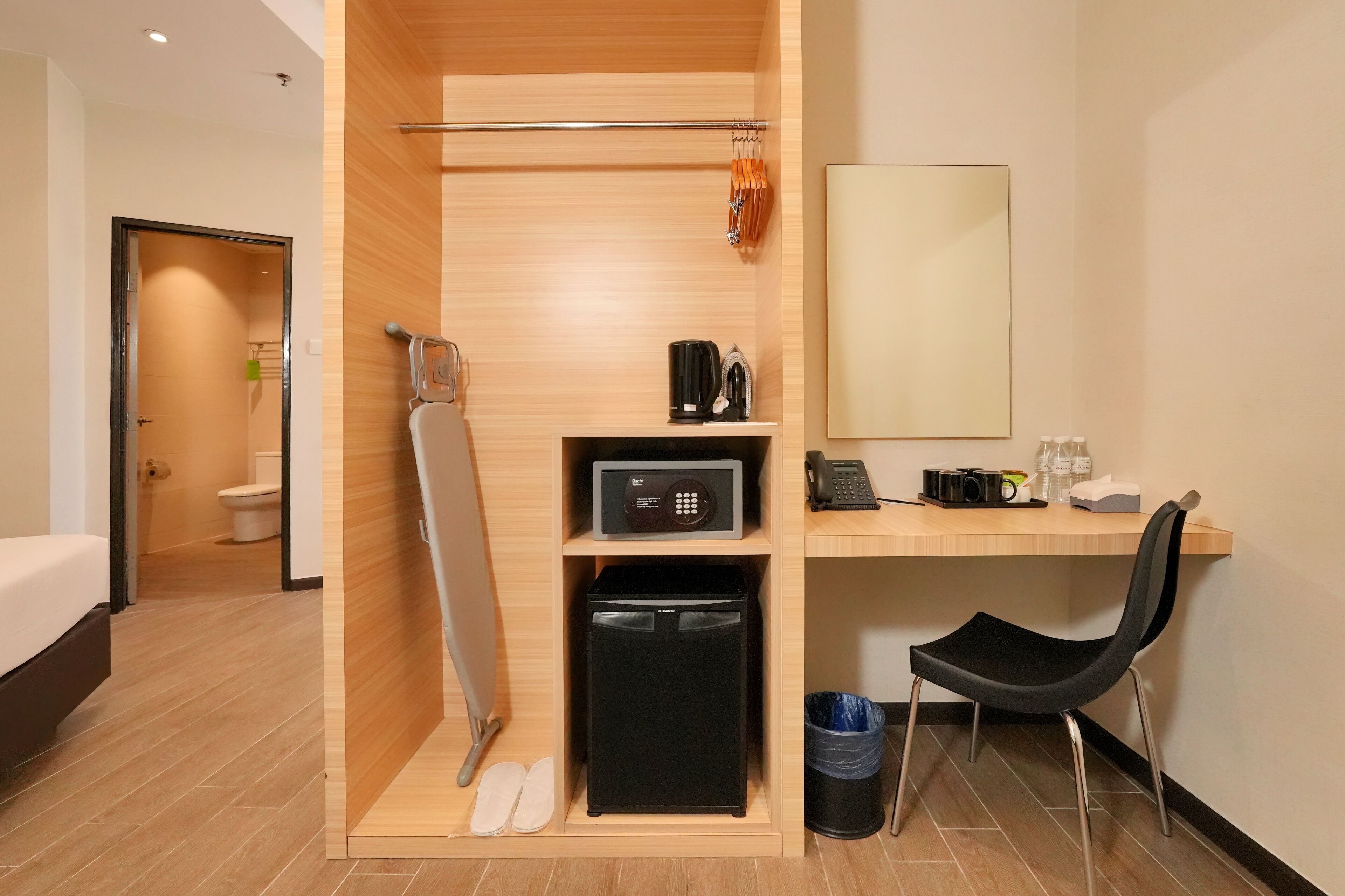 Una habitación de hotel compacta con un escritorio de madera, una silla negra, un refrigerador pequeño, un microondas, una cafetera y un cubo de basura.