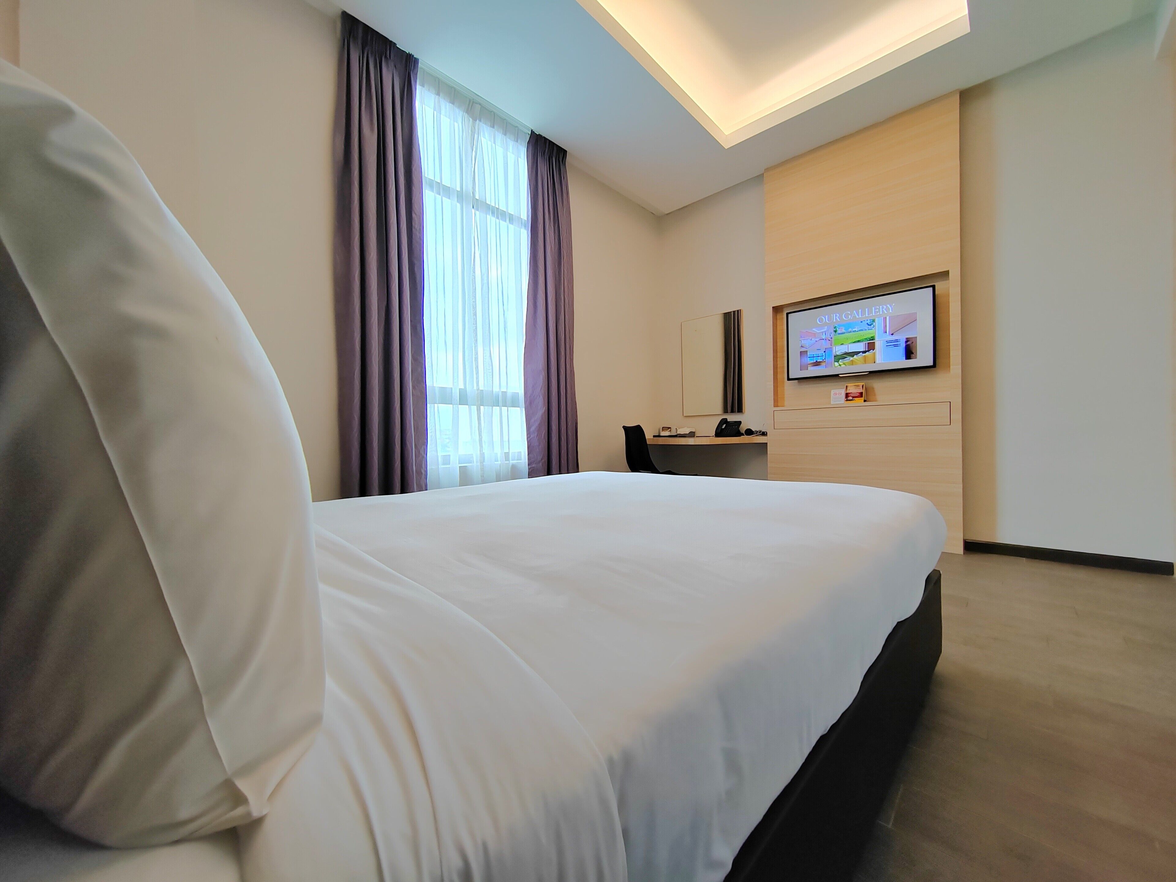 Habitación de hotel con una cama grande, un escritorio, una silla, un televisor en la pared y una ventana con cortinas.