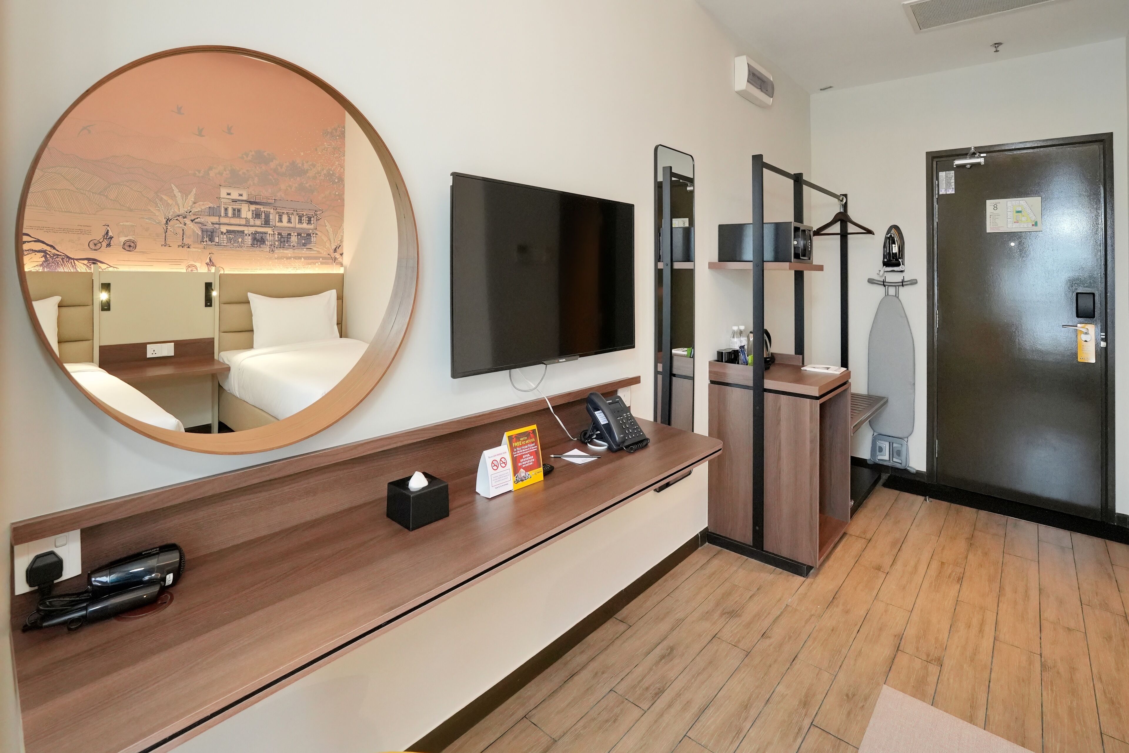 A hotel room with a large round mirror, a flat-screen TV, a wooden console table with a telephone, a small tissue box, and a sign.