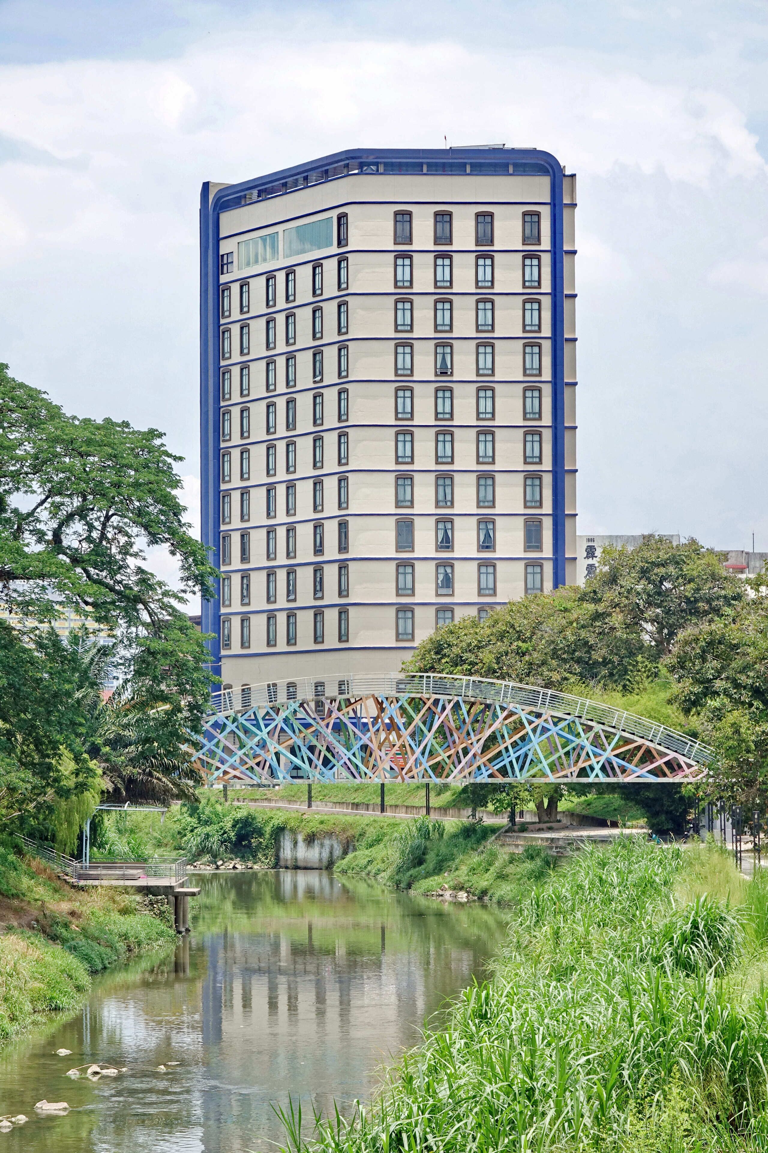 Un edificio alto de diseño moderno, un puente con estructura de celosía de colores y un río tranquilo en primer plano.