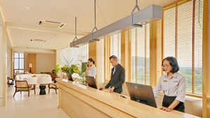 Un área de recepción con un mostrador de madera, dos empleados en sus puestos de trabajo y un espacio de espera con sillas y una mesa.