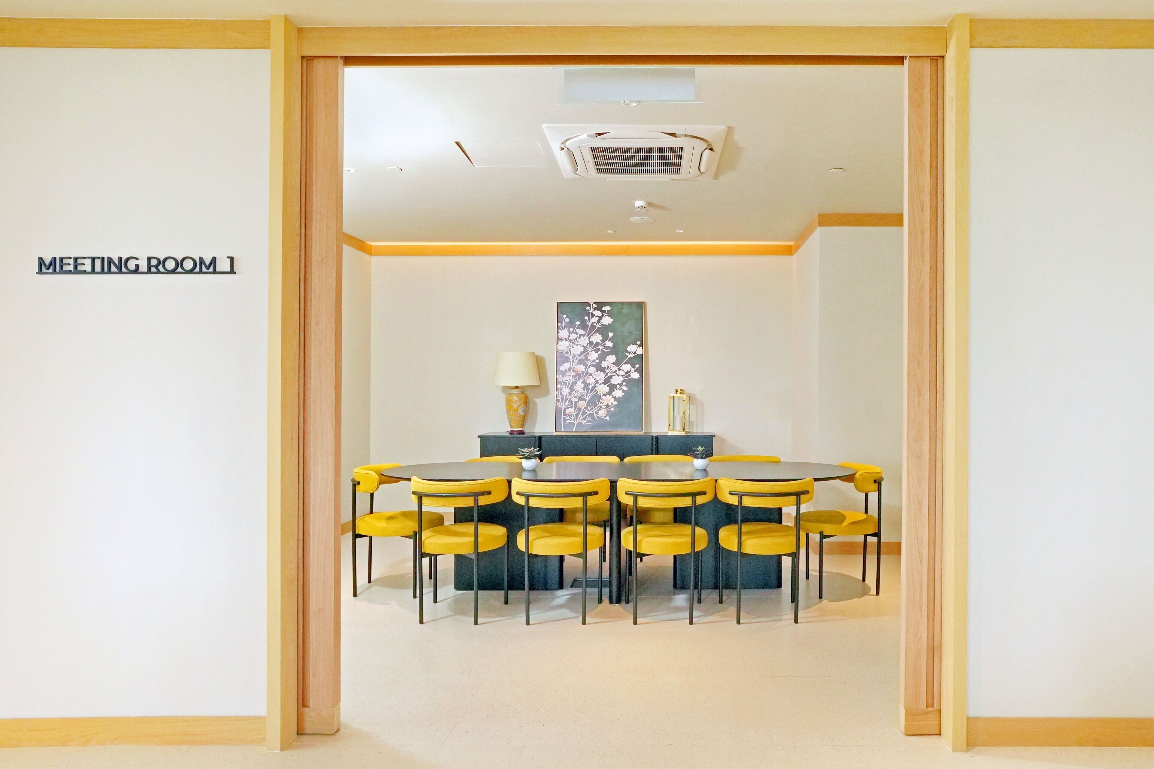Uma sala de reuniões com mesa, cadeiras, uma placa indicando Sala de Reuniões 1 e uma abertura no teto.