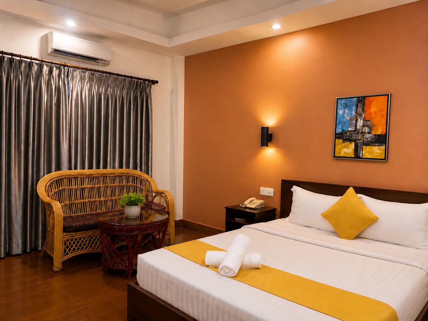 A hotel room with a bed, a chair, a small table with a plant, a nightstand with a phone, and a painting on the wall.