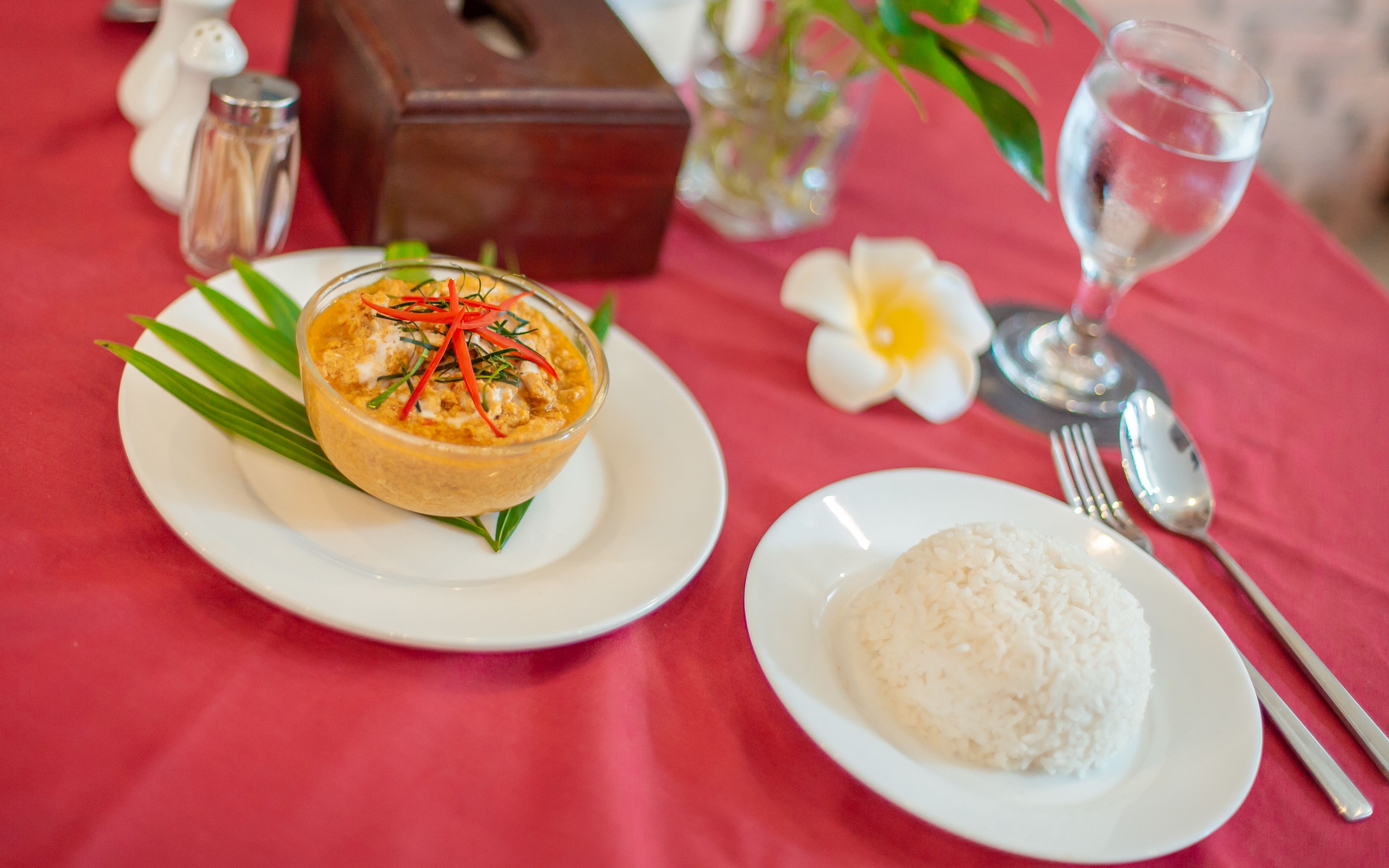 A table with a red tablecloth, a white plate with a bowl of food, a white plate with rice, a glass of water, and a flower.
