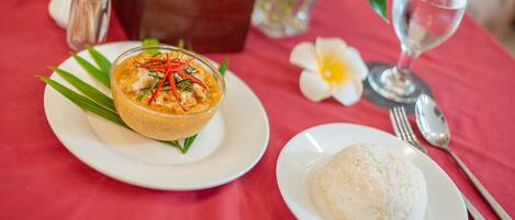 A table with a red tablecloth, a white plate with a bowl of food, a white plate with rice, a glass of water, and a flower.
