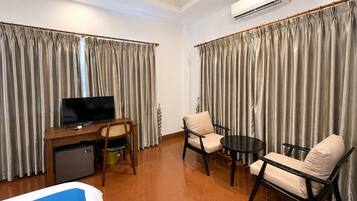 A hotel room with a bed, a desk with a television, a small refrigerator, two armchairs, a coffee table, and curtains.