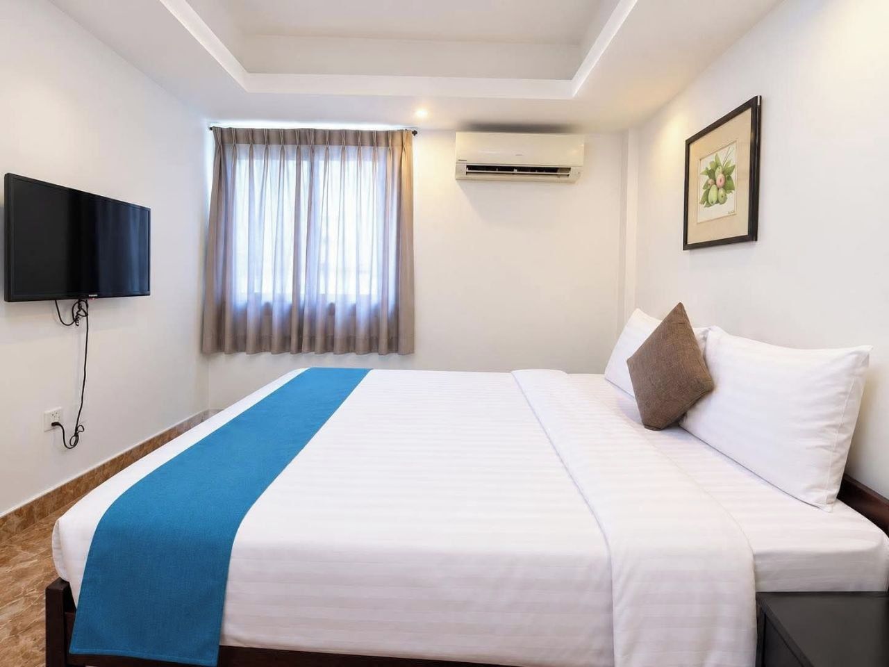 A hotel room with a bed, a flat-screen TV, an air conditioning unit, a window with curtains, and a framed picture on the wall.