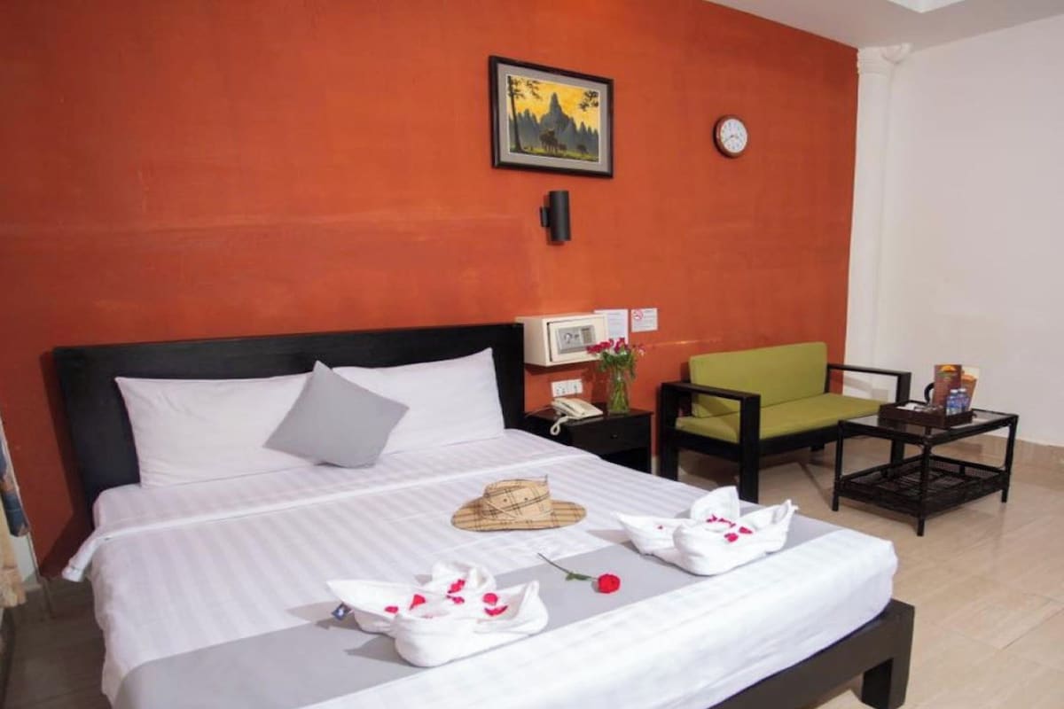 A hotel room with a bed, a bedside table with a clock, a chair, and a small table with a book.