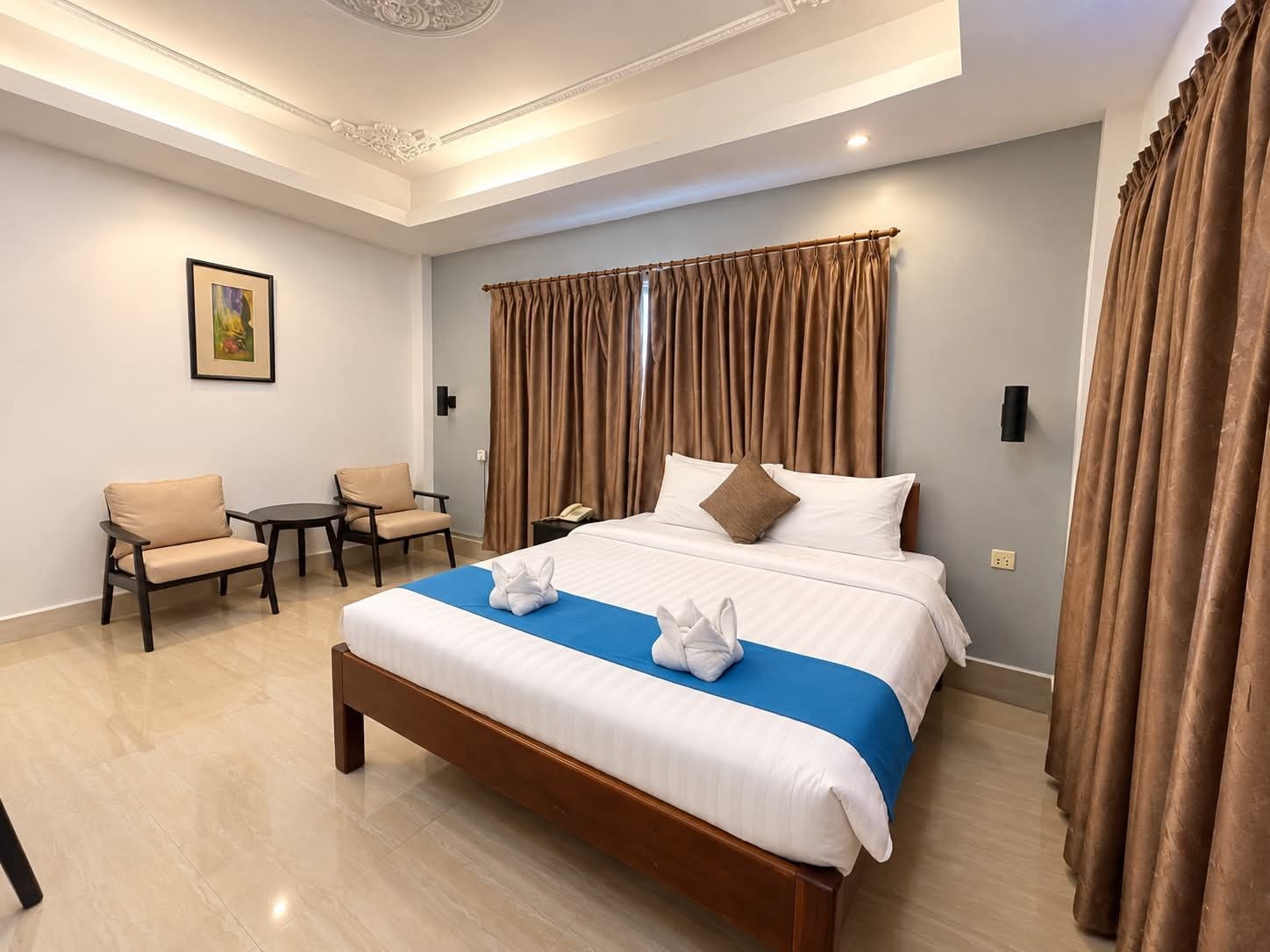 A hotel room with a large bed, two chairs, a small table, and a painting on the wall.