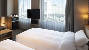 Quarto de hotel com uma cama grande, televisão na parede, escrivaninha com luminária e vista da cidade através de amplas janelas.