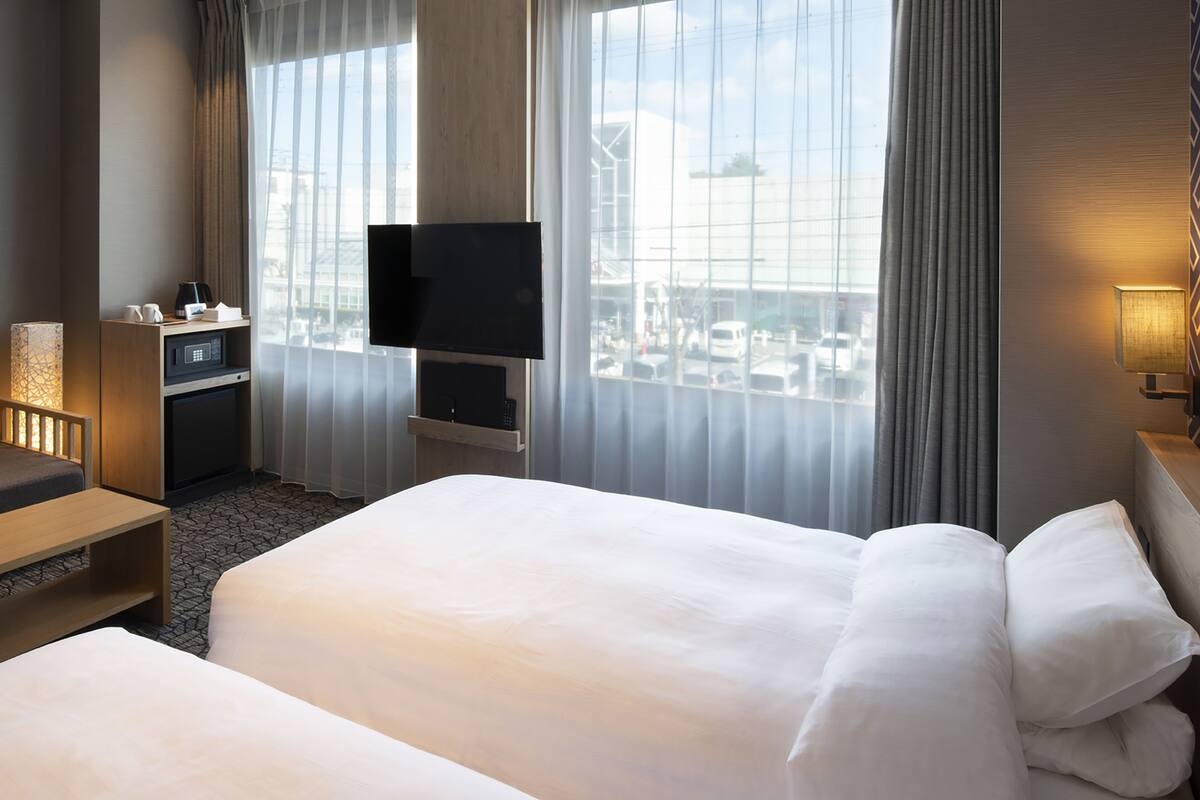 A hotel room with a large bed, a TV mounted on the wall, a desk with a lamp, and a view of the city through large windows.
