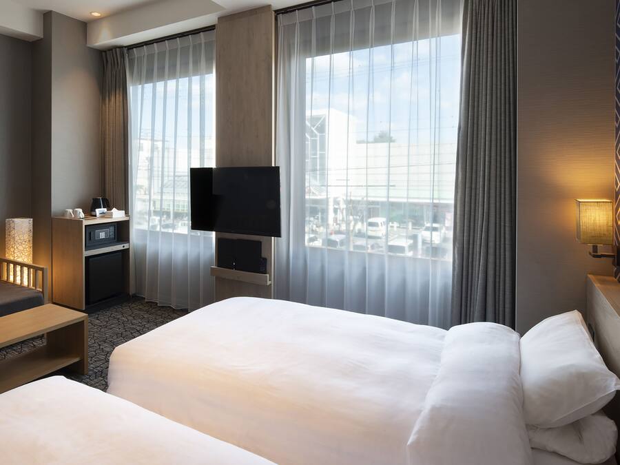 Une chambre d’hôtel dotée d’un grand lit, d’un téléviseur fixé au mur, d’un bureau avec une lampe et offrant une vue sur la ville grâce à de grandes fenêtres.