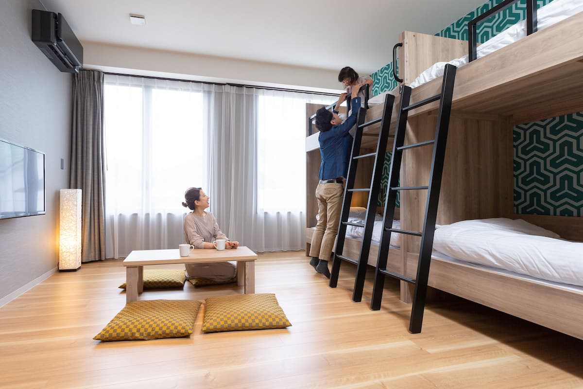 A hotel room with a bunk bed, a desk with two chairs, a standing lamp, and a large window with curtains.