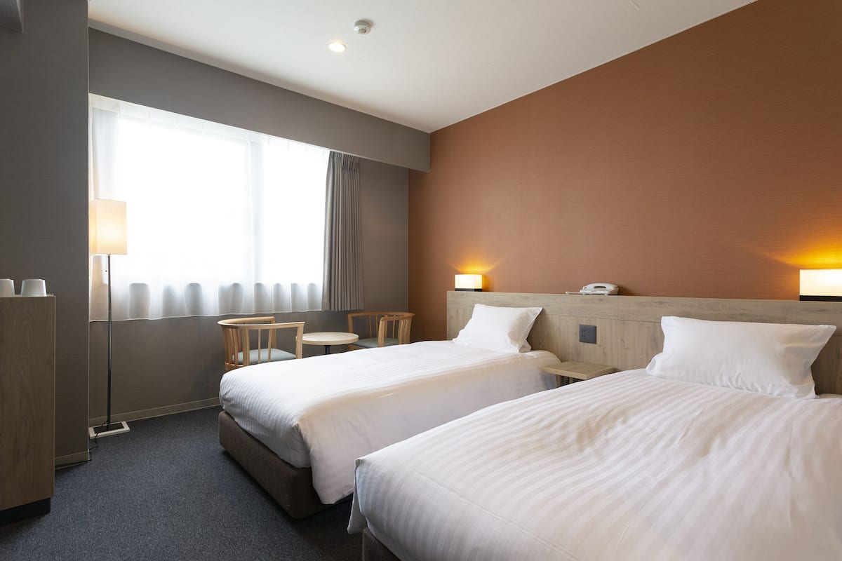 A hotel room with two beds, a desk, a chair, a lamp, and a window with curtains.