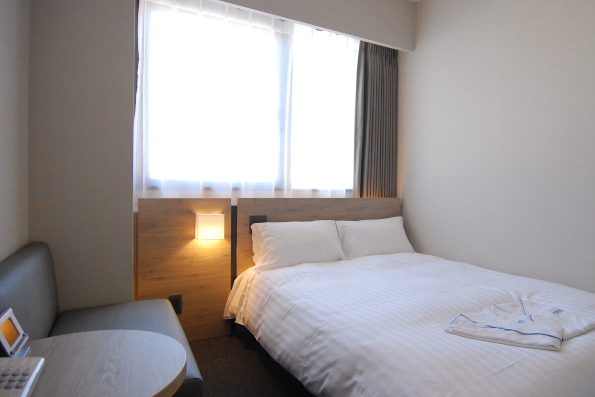 A hotel room with a bed, a bedside table, a lamp, and a window with curtains.