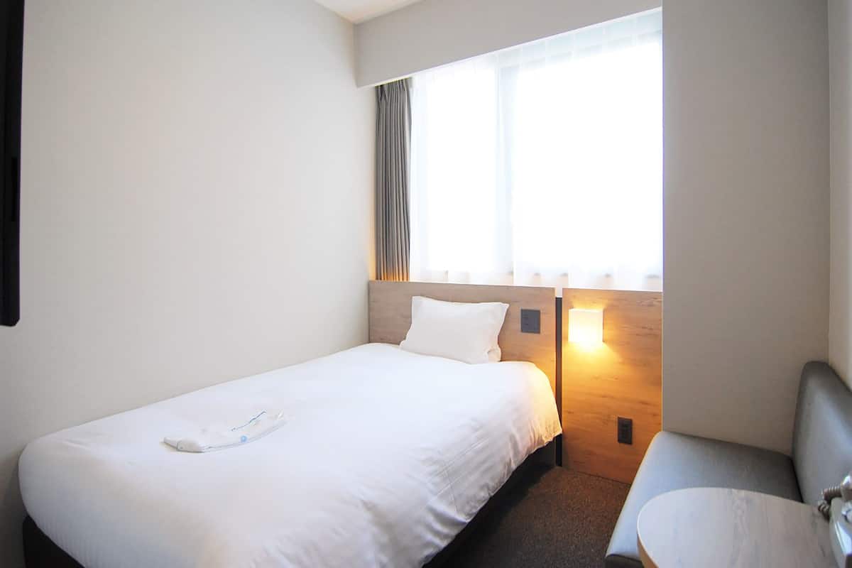A hotel room with a single bed, a small bedside table, a chair, and a window with curtains.