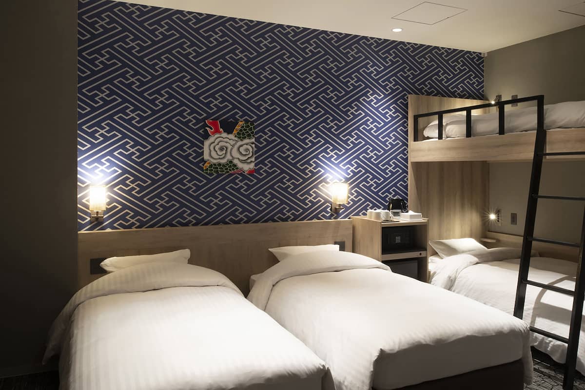 A hotel room with two beds, a bunk bed, a patterned wall, and two bedside lamps.