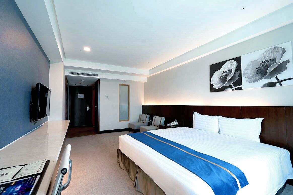 A hotel room with a large bed, a desk with a computer, a TV, and artwork on the wall.