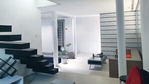 Un interior moderno con una escalera que conduce a un segundo piso, una zona de estar central con una mesita de café y una planta en maceta.