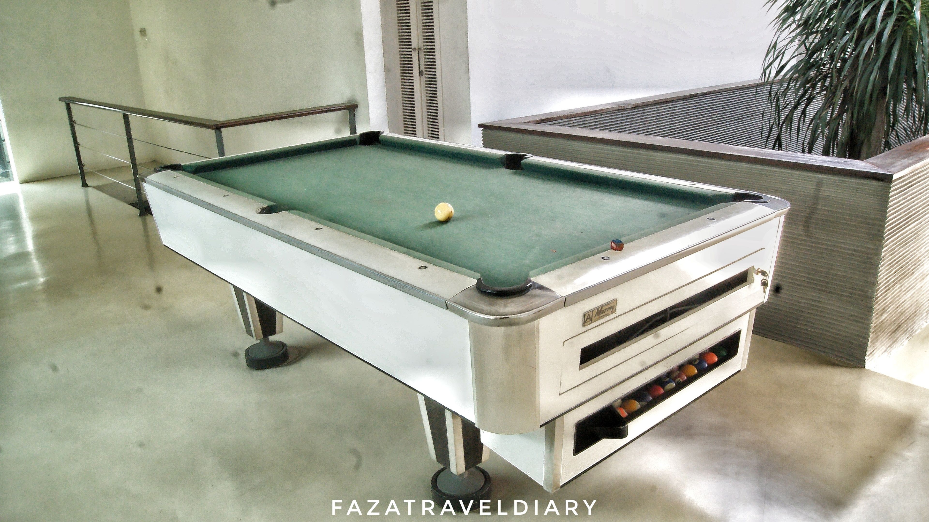 A pool table with a cue ball in the center, a white cabinet with a slot for balls, and a metal railing.