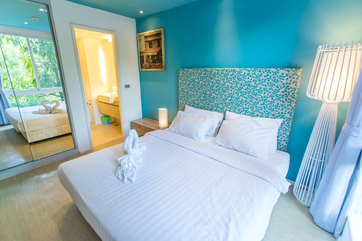 A hotel room with a large bed, a mirror, a bathroom, and a view of greenery.