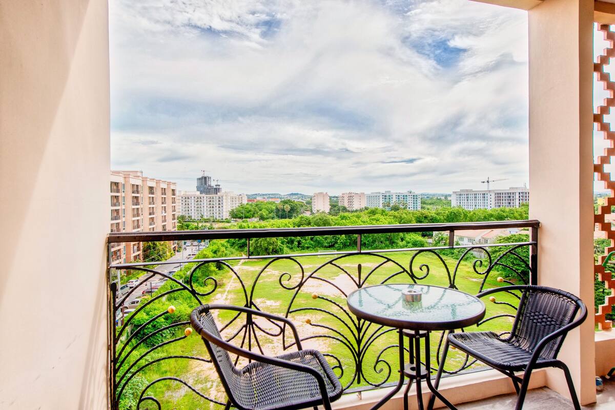 A balcony with two wicker chairs and a glass table, overlooking a cityscape with buildings and greenery.