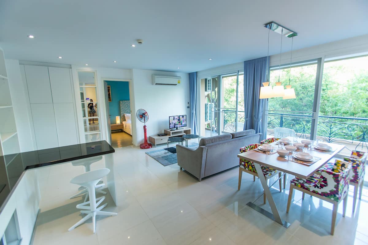 A modern living room with a sofa, dining table, and bar stools. There is a TV, a red fan, and a balcony with a view of greenery.