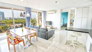 A modern living room with a sofa, a dining table with colorful chairs, a kitchen area with a bar stool, and a large window with a city view. - Atlantis Residence - Huge Saltwater Waterpark, Gym & Kids Playground - Free Parking (Pattaya)