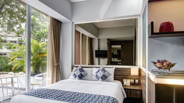 A hotel room with a large bed, a bedside table with a lamp, a TV mounted on the wall, and a view of greenery outside.