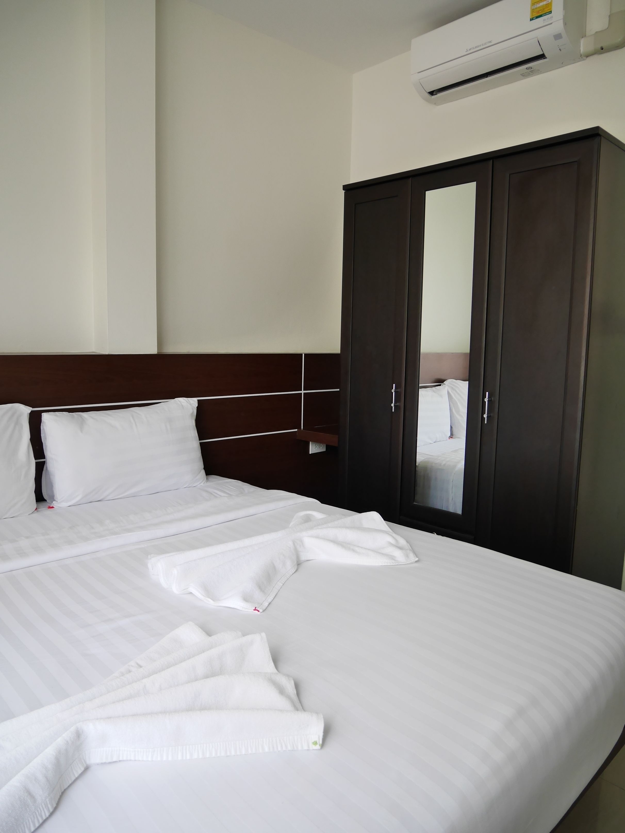 A neatly made bed with white linens and a folded towel, a dark wooden wardrobe, and a wall-mounted air conditioning unit.